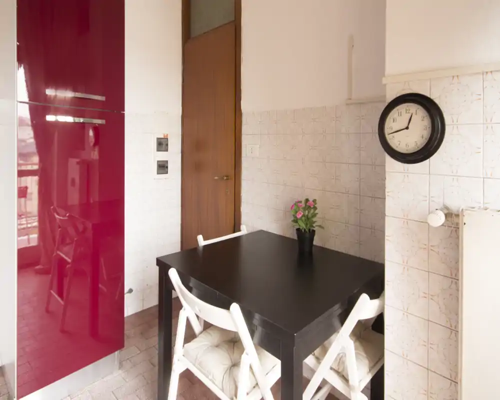 Angolo pranzo compatto con tavolo in legno scuro, quattro sedie pieghevoli bianche con cuscini e una piccola pianta in vaso; luce naturale a sinistra e piastrelle calde alle pareti rendono l'ambiente accogliente e funzionale.