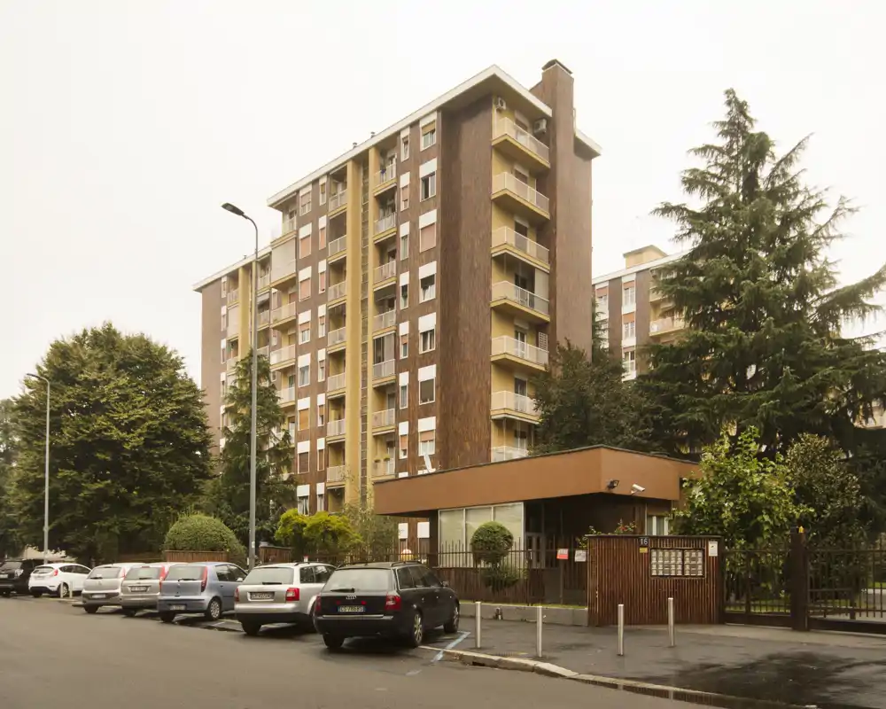 Vista esterna di un edificio residenziale multi-piano con auto parcheggiate lungo la strada e alberi maturi che inquadrano la proprietà.