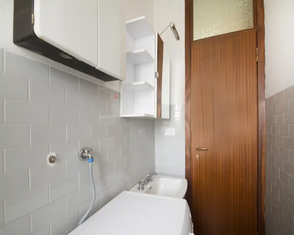 Piccola area bagno piastrellata con lavandino, pensili a muro e attacco per lavatrice; luminosa, pulita e arredamento essenziale.