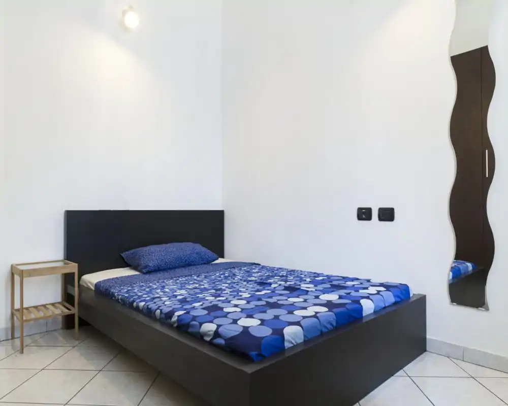 Camera da letto minimal e ben illuminata con un letto matrimoniale basso in stile moderno, completo di biancheria blu a motivo e un piccolo comodino. Pareti bianche pulite e uno specchio ondulato decorativo rendono l'ambiente contemporaneo e ordinato.
