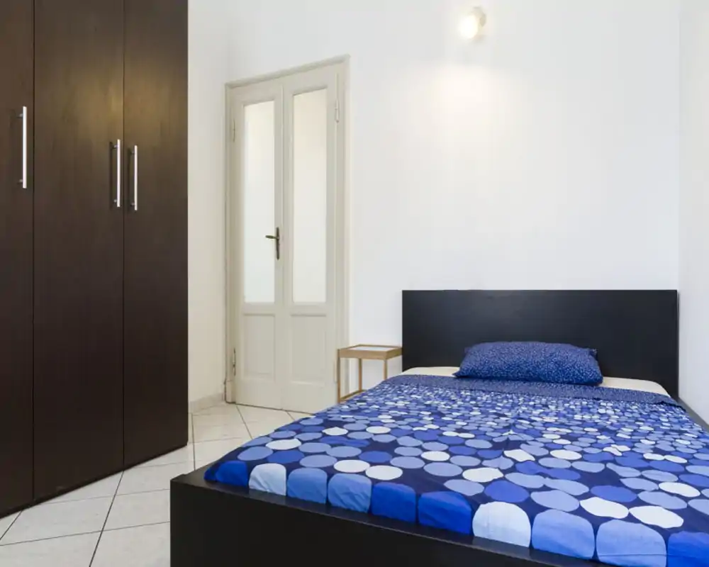 Camera da letto luminosa e dal design essenziale con letto matrimoniale, testiera scura e armadio alto color cioccolato. Pareti bianche e pavimento piastrellato creano un ambiente arioso e pulito.