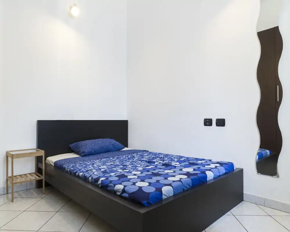 Camera da letto semplice e moderna con un letto basso scuro, copripiumino blu a fantasia e un cuscino abbinato; mobilio minimo. Pareti bianche e uno specchio alto conferiscono luminosità e senso di spazio.