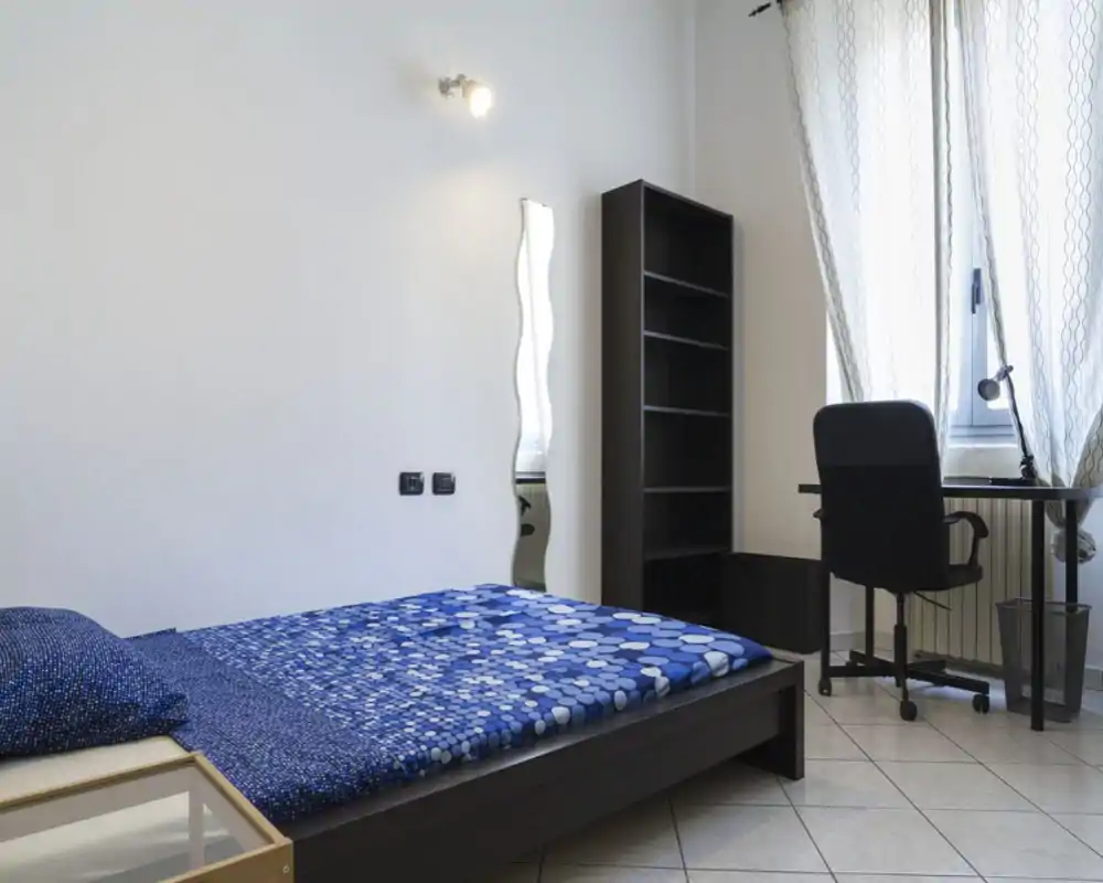 Camera da letto moderna e luminosa con letto matrimoniale con copriletto blu a fantasia, una libreria a giorno e una scrivania vicino alla finestra — pulita, minimal e ben illuminata per annunci.