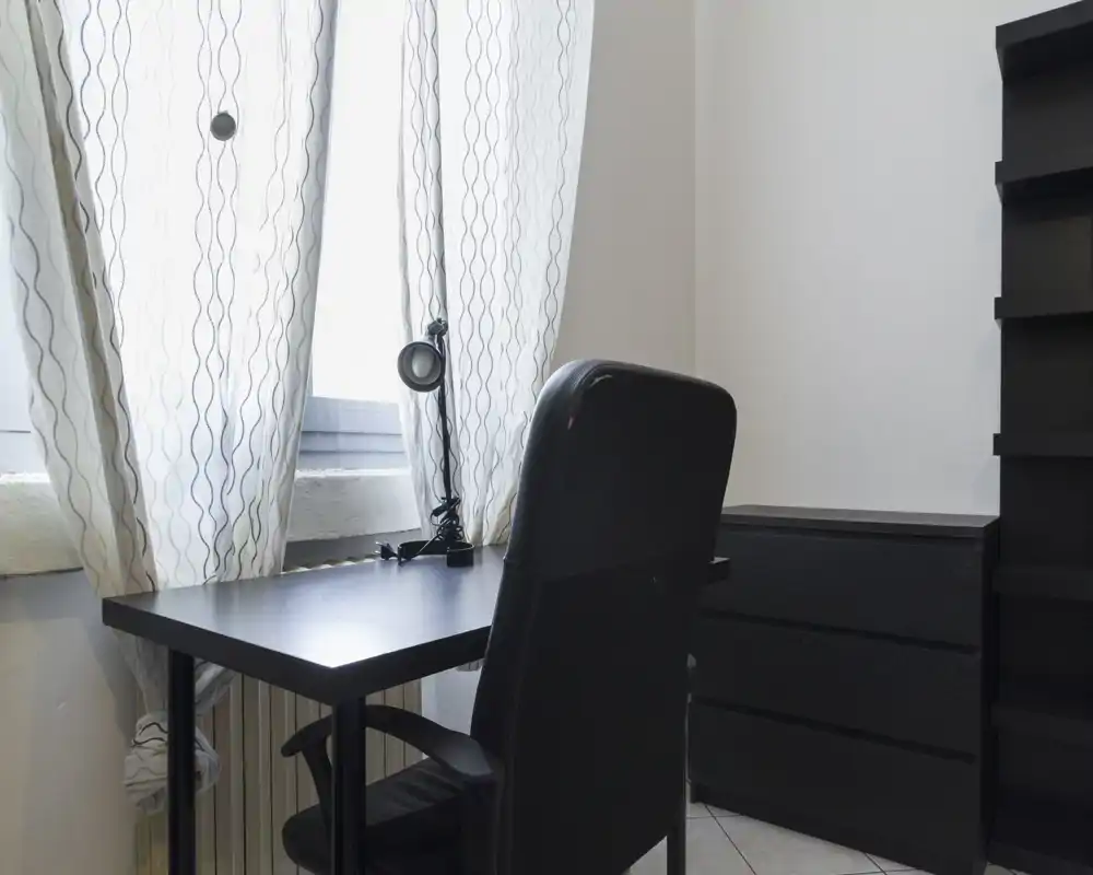 Angolo di lavoro luminoso e minimal con scrivania nera, sedia ergonomica e lampada da tavolo accanto a una finestra con tende — ideale per studio o smart working.