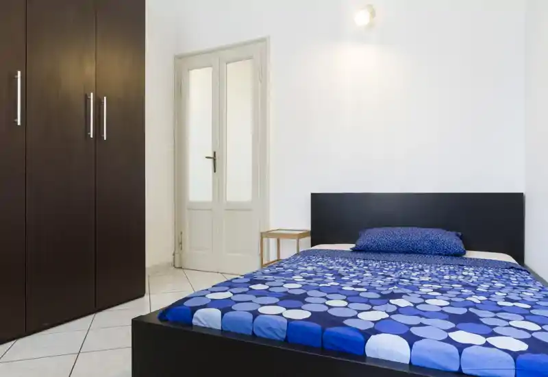 Camera da letto luminosa e minimal con letto matrimoniale con biancheria blu a motivi, testiera scura e grande armadio — linee pulite e arredamento semplice, ideale per affitti brevi.