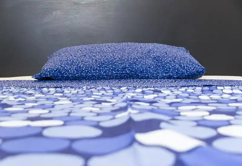 Foto luminosa e ben composta della camera da letto, con un letto rifatto in primo piano con biancheria blu a motivi e cuscino coordinato contro una testata scura.