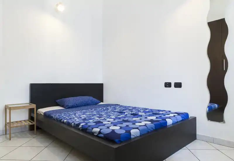 Camera da letto luminosa e minimal con letto basso scuro e biancheria blu a motivi, piccolo comodino e specchio a figura intera ondulato — spazio pulito, moderno e ben illuminato.