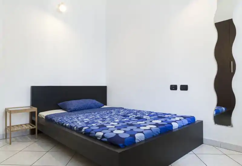 Dormitorio minimalista y bien iluminado con una cama baja oscura y ropa de cama azul estampada; mesita sencilla y espejo ondulado en la pared.