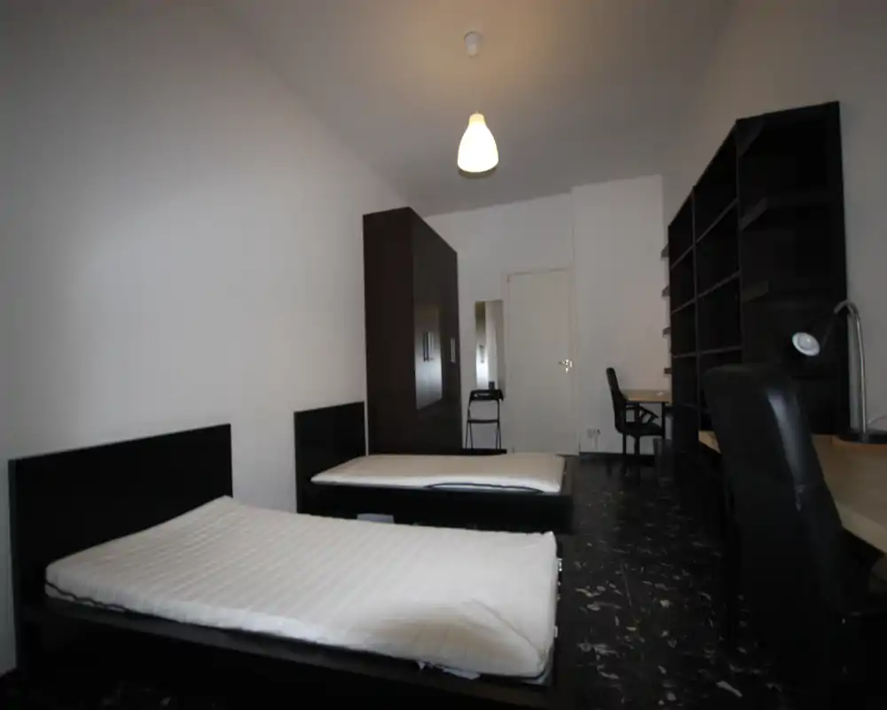 Amplia habitación con dos camas individuales, estructuras de madera oscura y mobiliario sencillo con armario y escritorios — luminosa, ordenada y adecuada para alojamiento compartido.