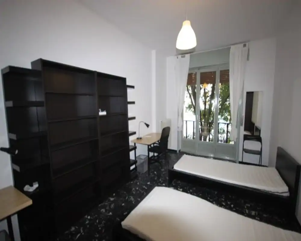 Amplia habitación doble con dos camas individuales, escritorio, estanterías y balcón que aporta luz natural. Mobiliario sencillo y suelo efecto mármol que crean un espacio limpio y funcional, ideal para estudiantes o compañeros de piso.