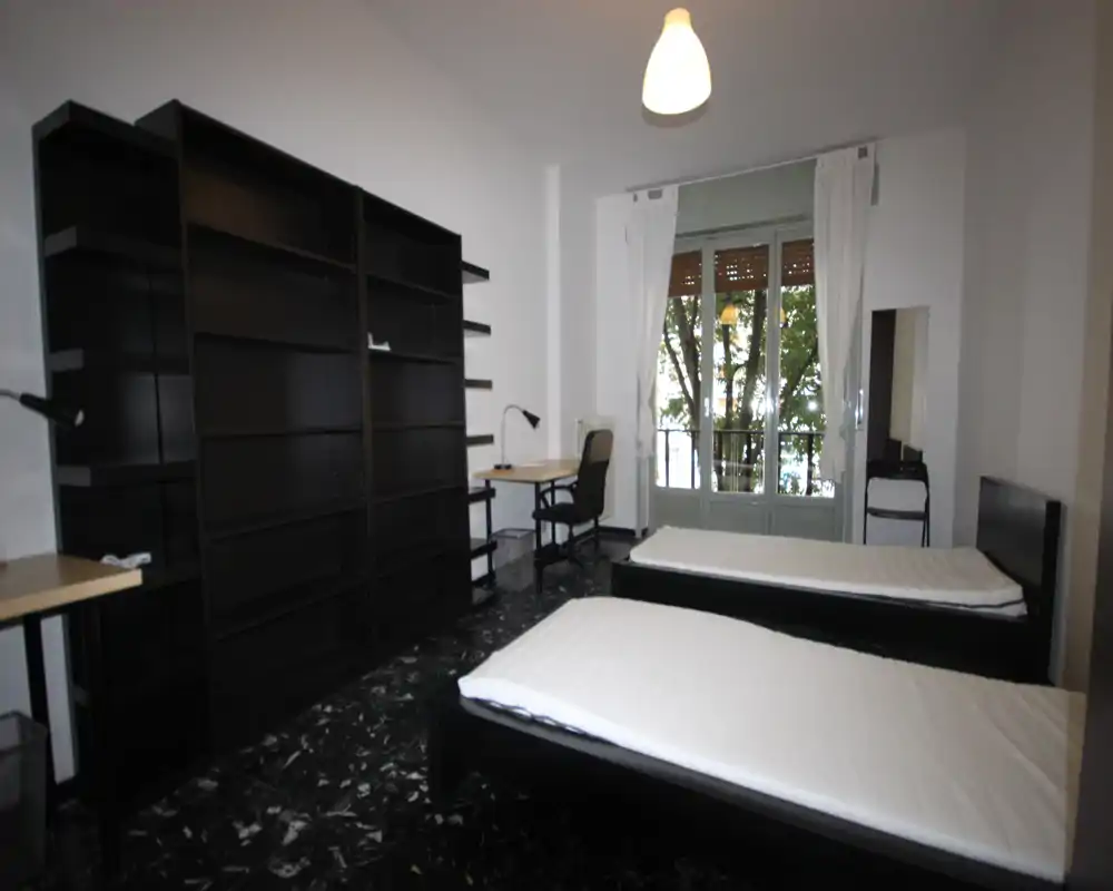 Amplia habitación doble con dos camas individuales, una gran estantería, escritorio y acceso al balcón que aporta luz natural. Mobiliario sencillo y funcional, ideal para estudiantes o alquiler compartido.