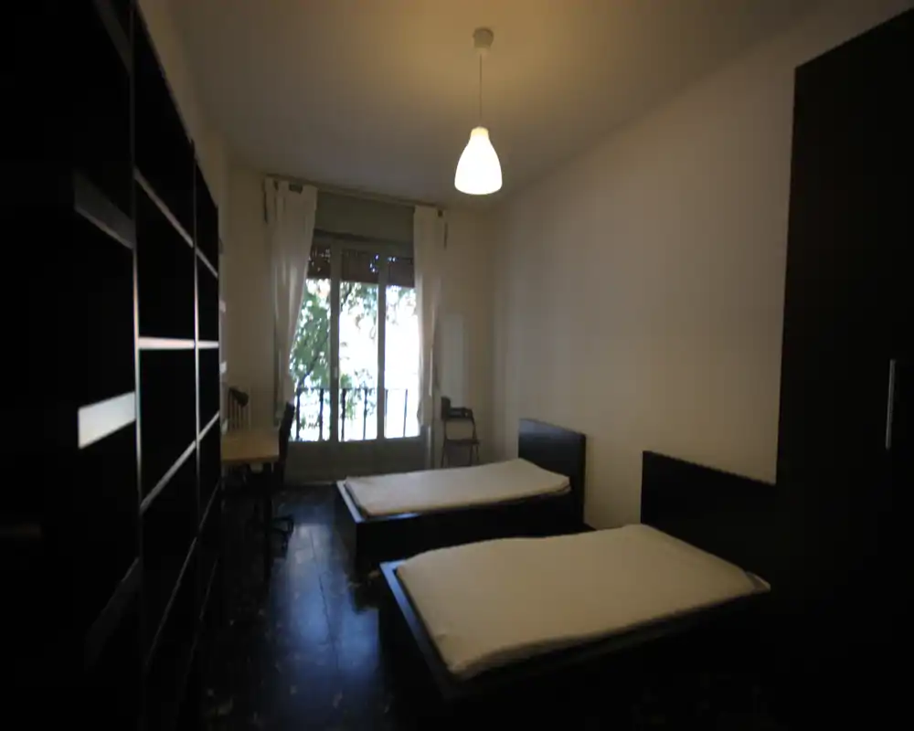 Dormitorio doble con dos camas individuales, amplia estantería, escritorio junto a la ventana y balcón que aporta luz natural — distribución sencilla y funcional, ideal para estudiantes o compañeros de piso.