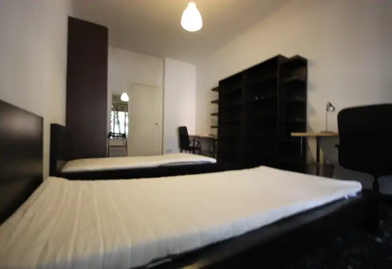 Dormitorio luminoso con dos camas individuales, mobiliario negro moderno y una gran estantería; zona de trabajo con mesa y silla al fondo. Distribución limpia y funcional, ideal para estudiantes o alquiler compartido.