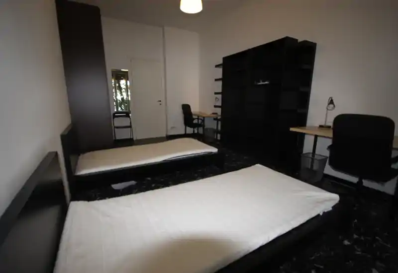 Dormitorio doble con dos camas individuales, mobiliario negro sencillo y dos zonas de estudio: distribución funcional y luminosa, perfecta para compañeros o estudiantes.