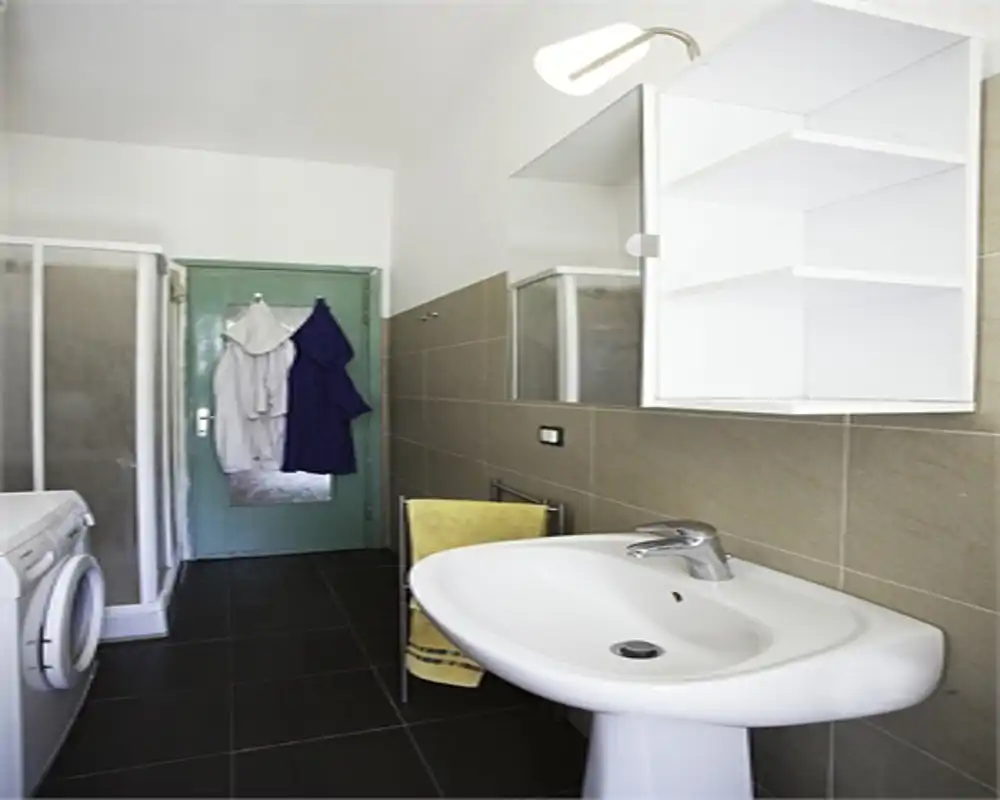 Baño limpio y moderno con lavabo pedestal, cabina de ducha, lavadora y armario mural; azulejos en tonos neutros y buena iluminación que hacen el espacio luminoso y funcional.