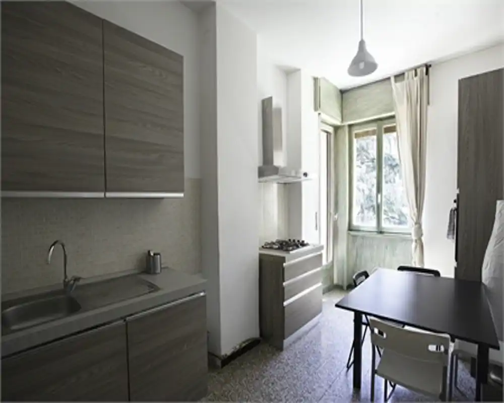 Cucina luminosa e moderna con pensili in legno grigio, lavello in acciaio e un piccolo tavolo da pranzo vicino alla finestra che illumina l'ambiente.