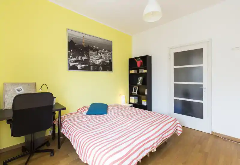 Camera da letto luminosa e ordinata con letto matrimoniale coperto da lenzuola a righe, una scrivania e una libreria. La parete gialla e il quadro aggiungono un tocco moderno e vivace.