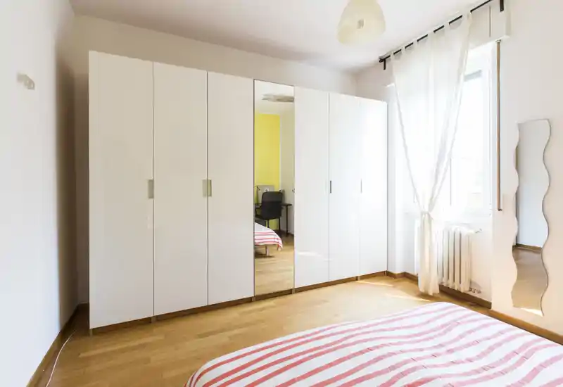 Camera da letto luminosa e semplice con copriletto a righe, grande armadio bianco con anta a specchio, pavimento in legno e finestra soleggiata con tende leggere.