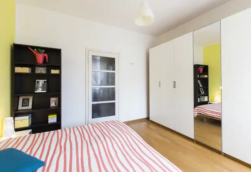 Camera da letto luminosa e moderna con letto a righe, grande armadio bianco con anta a specchio e una libreria nera; pavimento in legno e arredo semplice e fresco.