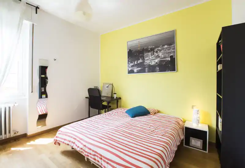 Camera da letto luminosa e moderna con letto matrimoniale, copriletto a righe, lampada da comodino e una piccola postazione di lavoro vicino alla finestra. Parete gialla e pavimento in legno danno un'atmosfera fresca e accogliente.