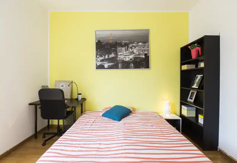 Camera da letto luminosa e ordinata con letto matrimoniale al centro contro una parete gialla, comodino con lampada e una scrivania su un lato. È presente una libreria alta per riporre oggetti e elementi decorativi, ideale per un ambiente accogliente e funzionale.