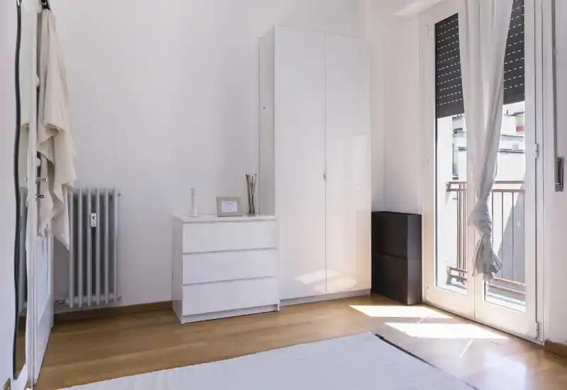 Rincón de dormitorio minimalista y luminoso con armario blanco y cómoda junto a la puerta del balcón que aporta abundante luz natural.