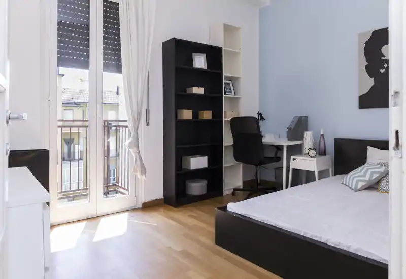 Dormitorio moderno y luminoso con cama doble, mesita de noche y un pequeño escritorio junto a la ventana que da al balcón. Tonos neutros y suelo de madera que generan un ambiente tranquilo y acogedor, perfecto para estudiantes o profesionales.