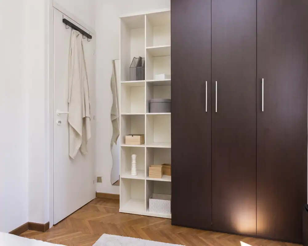 Angolo della camera ordinato con un ampio armadio in legno scuro, scaffalatura aperta e una porta con appendini; toni neutri e pavimento in parquet creano un'atmosfera minimal e tranquilla.