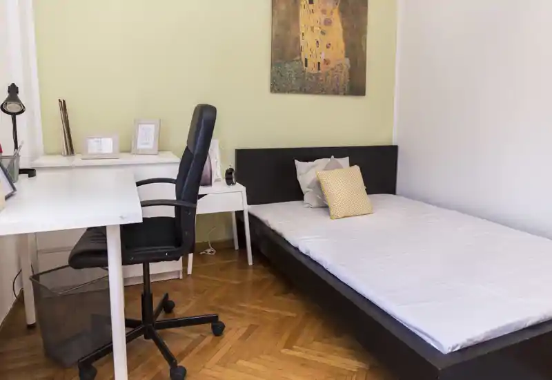 Chambre lumineuse avec un lit bas, une table de chevet simple et un bureau avec chaise pivotante. Parquet propre et un tableau accroché qui donnent un espace net et fonctionnel, idéal pour un étudiant ou une personne seule.