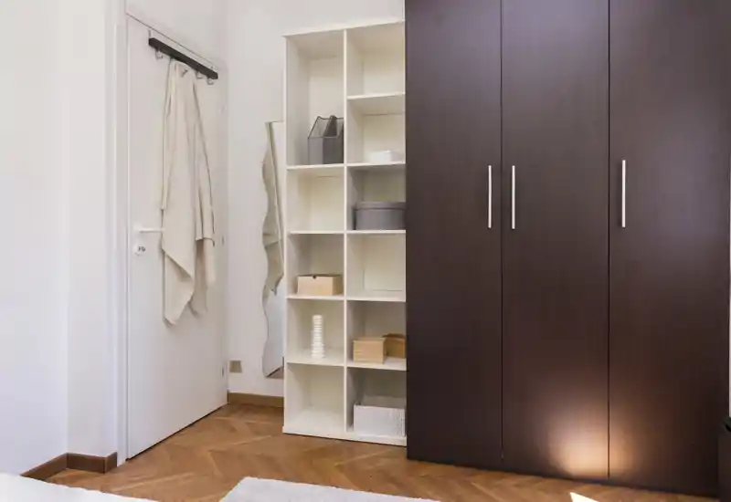Coin chambre soigné avec une grande armoire en bois foncé, des étagères ouvertes et une porte avec crochets ; tons neutres et parquet pour une atmosphère calme et minimaliste.