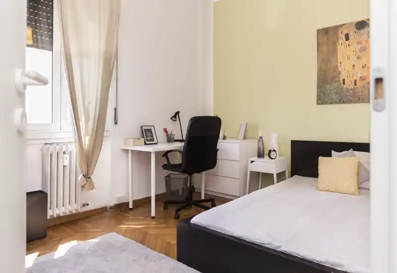 Chambre simple lumineuse et bien mise en scène avec lit rangé, table de chevet et un petit bureau près de la fenêtre — idéale pour étudiant ou professionnel seul.