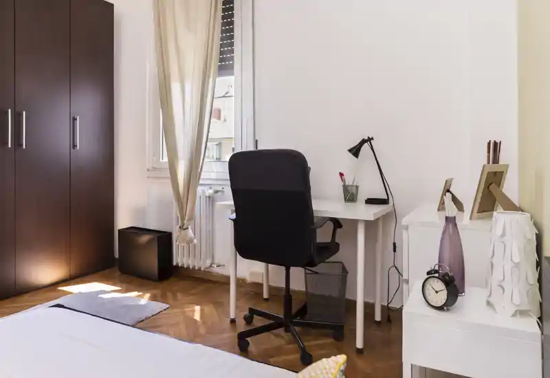 Chambre lumineuse et bien aménagée avec un coin de lit visible, un bureau et une armoire près de la fenêtre — idéale pour les locations courte durée recherchant un espace nuit propre et fonctionnel.