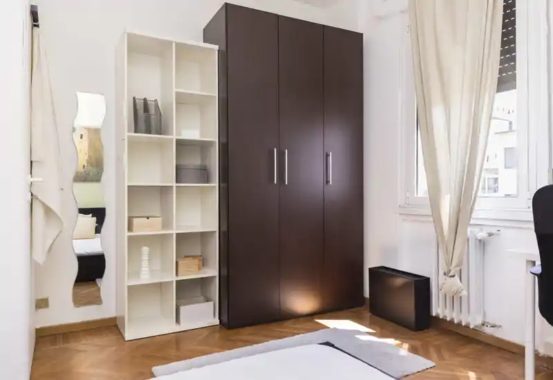 Coin de chambre lumineux et soigné avec le bord du lit visible, grand placard en bois foncé et étagères — la fenêtre ensoleillée apporte de la chaleur.