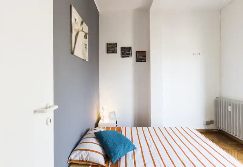 Chambre lumineuse et moderne avec un lit double rayé contre un mur gris, une lampe de chevet et un coussin turquoise pour une touche de couleur.