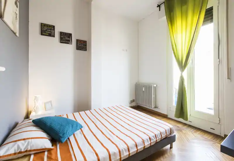 Chambre lumineuse et soignée avec un lit double aux draps à rayures orange, un coussin bleu sarcelle et une grande fenêtre avec rideaux verts laissant entrer la lumière naturelle.