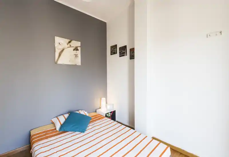 Camera da letto luminosa e ordinata con letto a righe, parete accentuata e lampada da comodino che creano un'atmosfera moderna e accogliente.