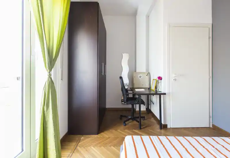 Coin de chambre lumineux et soigné avec lit rayé, bureau, armoire et grande fenêtre laissant entrer la lumière naturelle.