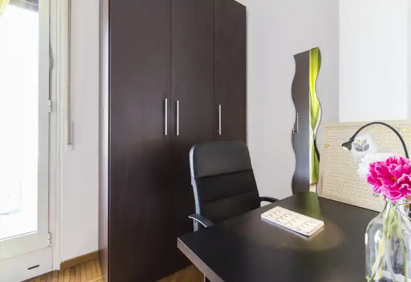 Coin salon/bureau compact avec armoire en bois foncé, bureau, fauteuil de bureau et lumière naturelle venant d'une porte; des fleurs fraîches apportent une touche accueillante.