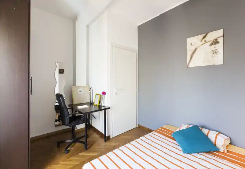 Chambre moderne et lumineuse avec lit rayé, coussin bleu-vert et un petit espace de travail. Parquet soigné et décoration neutre offrent une pièce calme, idéale pour se reposer et étudier.