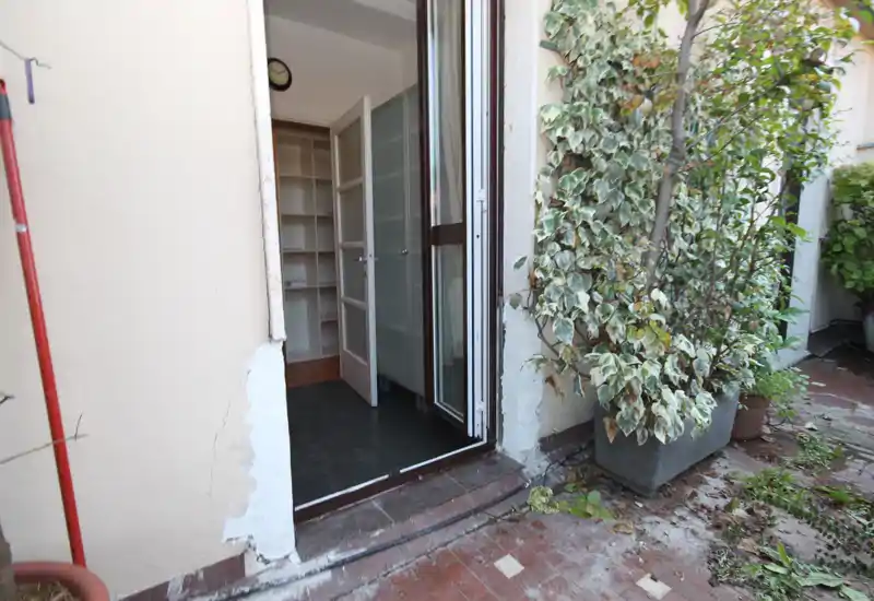 Petite terrasse privée avec plantes en pots et accès direct à l'appartement ; sol carrelé et porte donnant sur une pièce intérieure avec étagères.