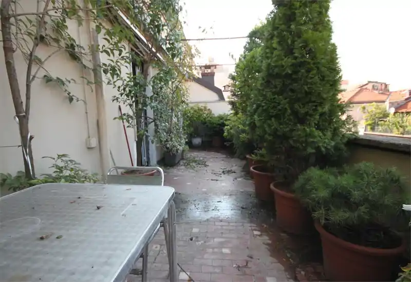 Terrasse étroite en ville avec sol carrelé, pots de conifères et arbustes le long du garde-corps et une table d'extérieur en métal au premier plan. Idéale pour un coin détente ou le jardinage en pot.