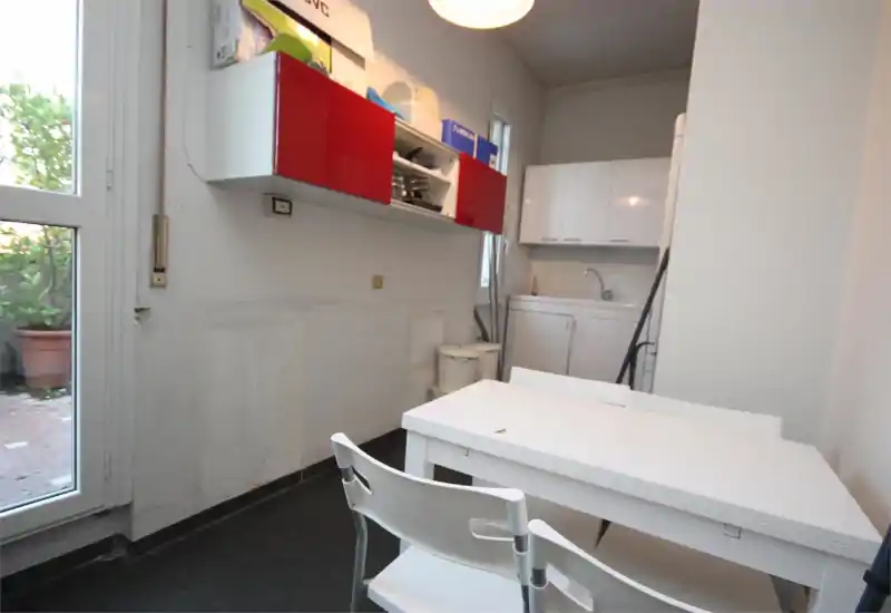 Cuisine compacte avec meubles blancs et placards muraux rouges, petit évier et table simple près d'une porte-fenêtre donnant sur l'extérieur.