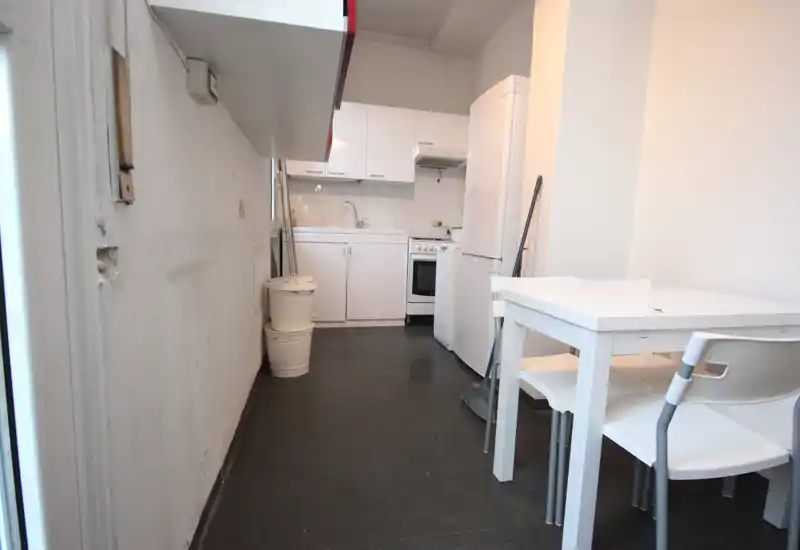 Petite cuisine équipée avec placards blancs, réfrigérateur et cuisinière compacts, ainsi qu'une table et chaises blanches simples — fonctionnelle et minimaliste.