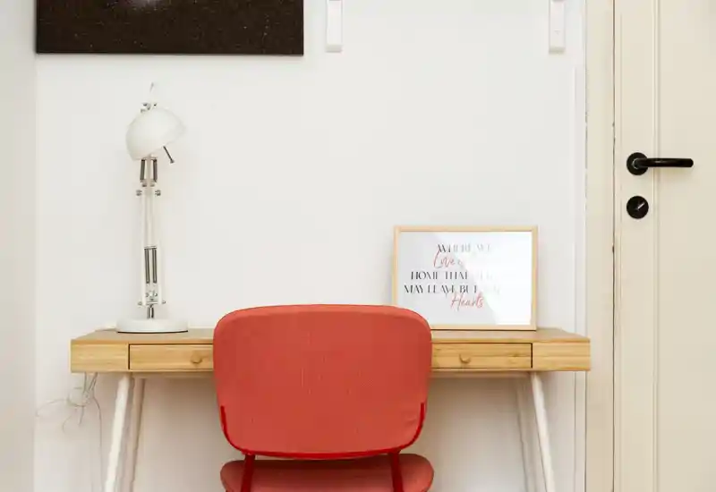 Compact workspace with a wooden desk, a white desk lamp and a framed print, paired with a red upholstered chair — bright and minimal setup ideal for a home office corner.