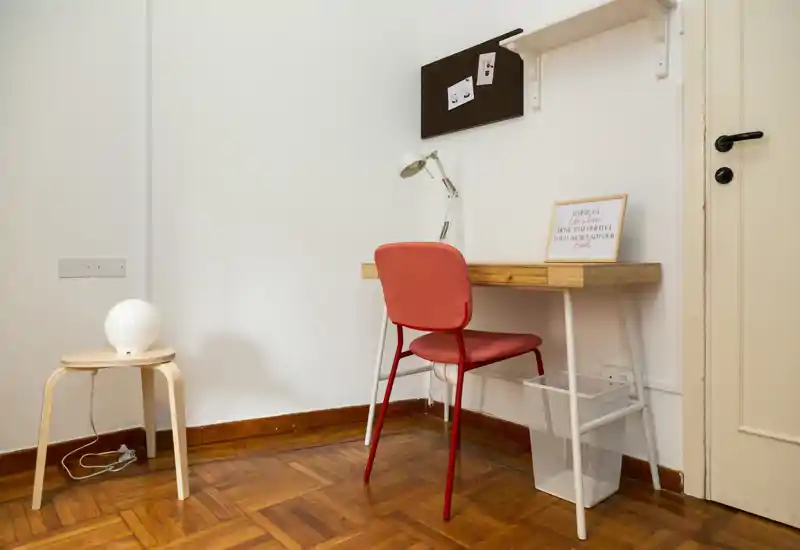 Compact, minimalist workspace in a room: wooden desk with a red chair, desk lamp, pinboard and a small side table on parquet flooring. Bright, tidy corner ideal for studying or remote work.
