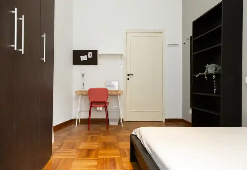 Dormitorio luminoso y ordenado con cama visible, suelo de parquet, un pequeño escritorio con silla roja y estantería abierta — ideal para una persona o pareja.