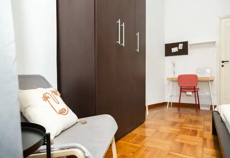 Bright, tidy bedroom corner with a visible wardrobe, a small desk and a partial view of the bed. Simple modern furnishings, wooden parquet floor and a cozy accent chair with decorative pillow.