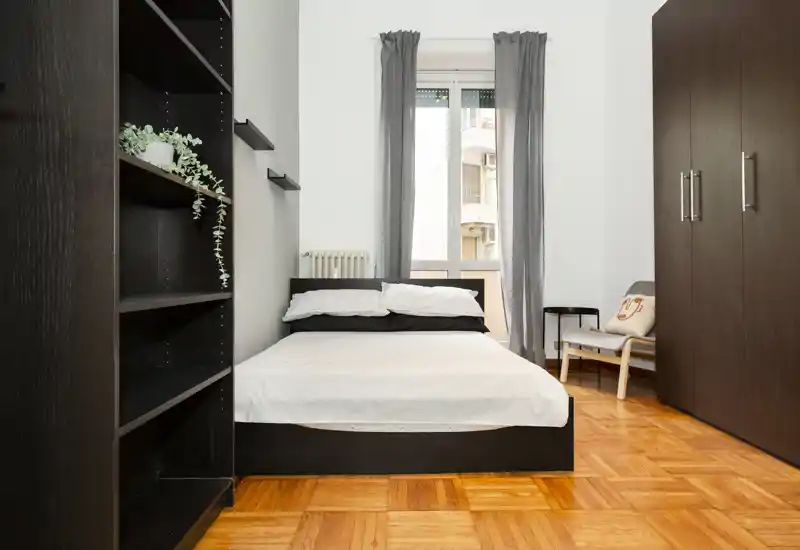 Dormitorio moderno y luminoso con cama doble centrada, armario alto y estanterías. La luz natural y el suelo de parquet aportan una sensación acogedora y minimalista, ideal para alquileres de corta estancia.