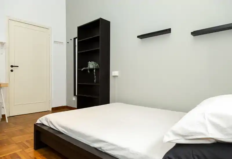 Dormitorio luminoso y de estilo sencillo con cama baja de madera oscura y ropa blanca, estantería alta junto a la puerta y decoración mínima.