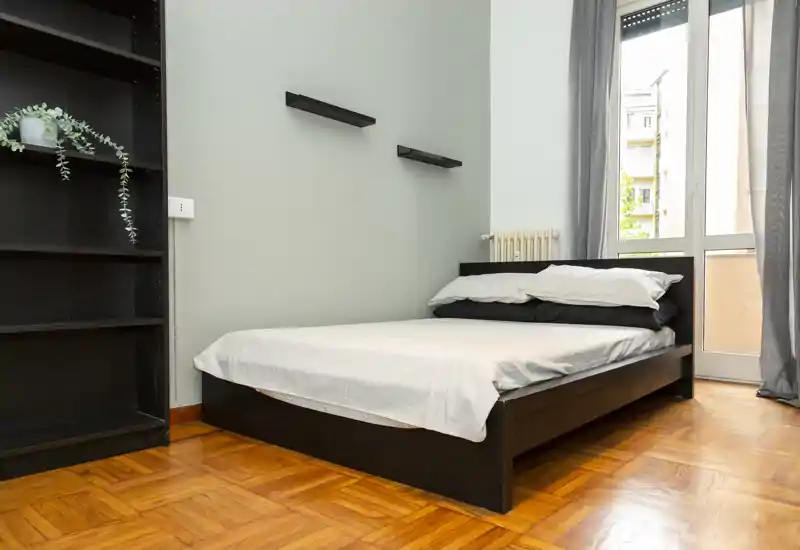 Dormitorio moderno y luminoso con cama doble baja de madera, ropa de cama blanca y una gran ventana que aporta luz natural. Estanterías sencillas y suelo de parquet crean un ambiente limpio y acogedor, perfecto para anuncios de alquiler.